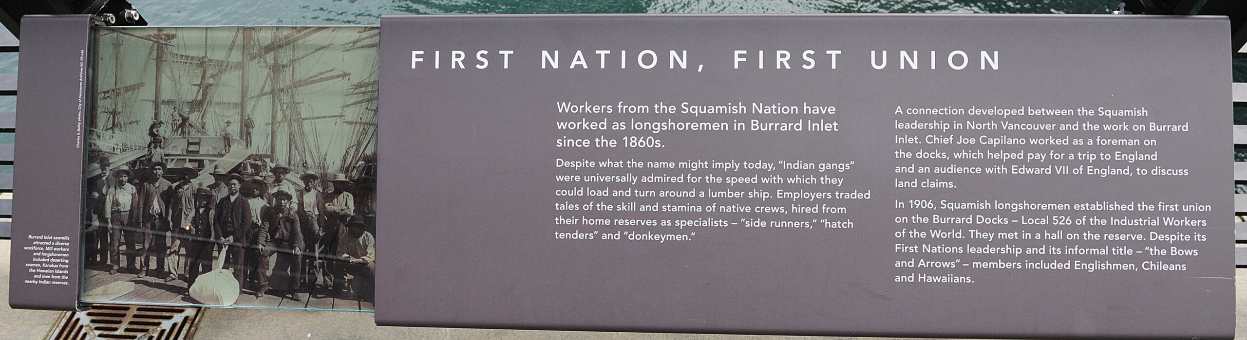 The plaque reads: Workers from the Squamish Nation have worked as longshoremen in Burrard Inlet since the 1860s. Despite what the name might imply today, “Indian gangs” were universally admired for the speed with which they could load and turn around a lumber ship. Employers traded tales of the skill and stamina of native crews, hired from their home reserves as specialists – “side runners,” “hatch tenders” and “donkeymen.” A connection developed between the Squamish leadership in North Vancouver and the work on Burrard Inlet. Chief Joe Capilano worked as a foreman on the docks, which helped pay for a trip to England and an audience with Edward VII of England, to discuss land claims. In 1906, Squamish longshoremen established the first union on the Burrard Docks – Local 526 of the Industrial Workers of the World. They met in a hall on the reserve. Despite its First Nations leadership and its informal title – “the Bows and Arrows” – members included Englishmen, Chileans and Hawaiians.