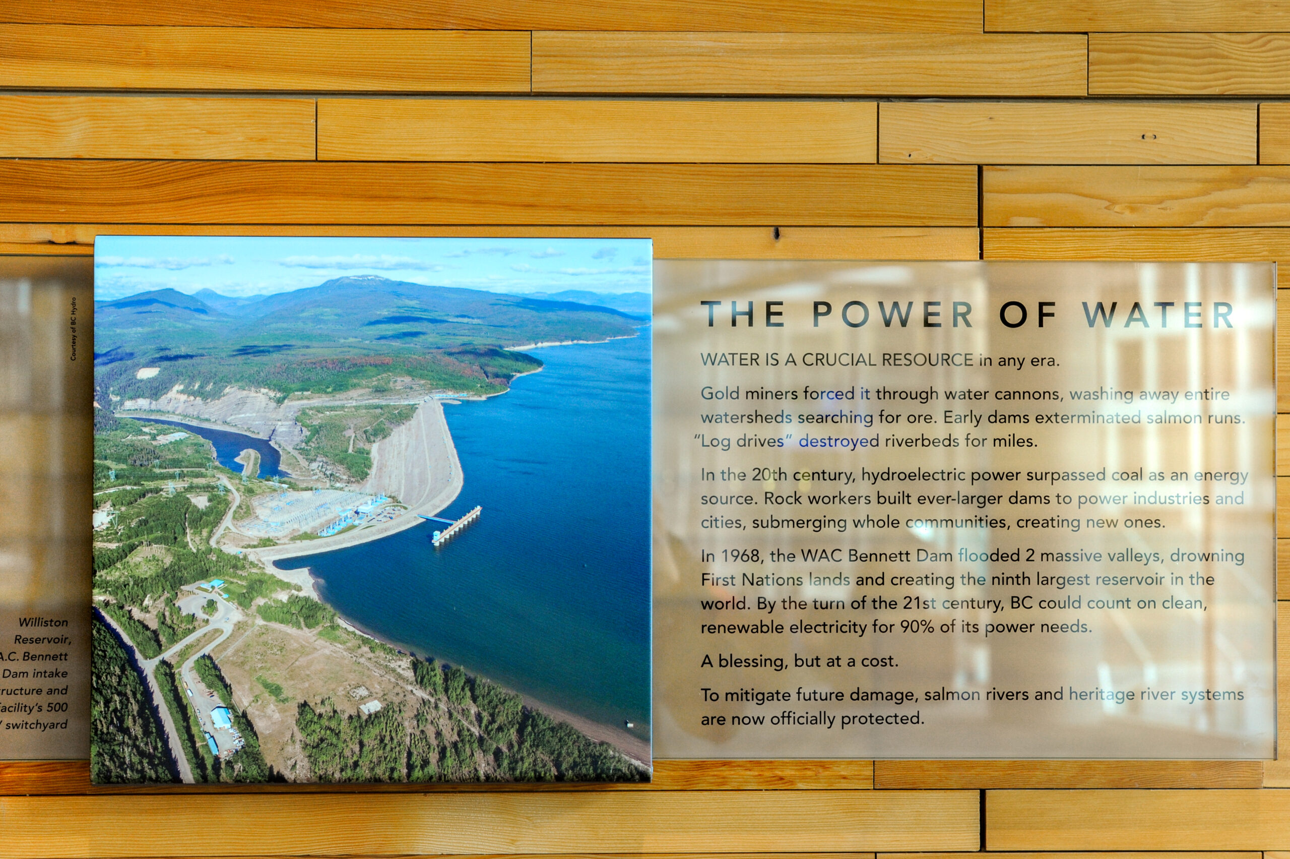 The plaque reads: Water is a crucial resource in any era. Gold miners forced it through water cannons, washing away entire watersheds searching for ore. Early dams exterminated salmon runs. Log drives destroyed riverbeds for miles. In the 20th century, hydroelectric power surpassed coal as an energy source. Rock workers built ever-larger dams to power industries and cities, submerging whole communities, creating new ones. In 1968, the WAC Bennett Dam flooded two massive valleys, drowning First Nations land and creating the ninth-largest reservoir in the world. By the turn of the 21st century, BC could count on clean, renewable energy for 90% of its power needs. A blessing, but at a cost. To mitigate further damage, salmon rivers and heritage river systems are now officially protected.