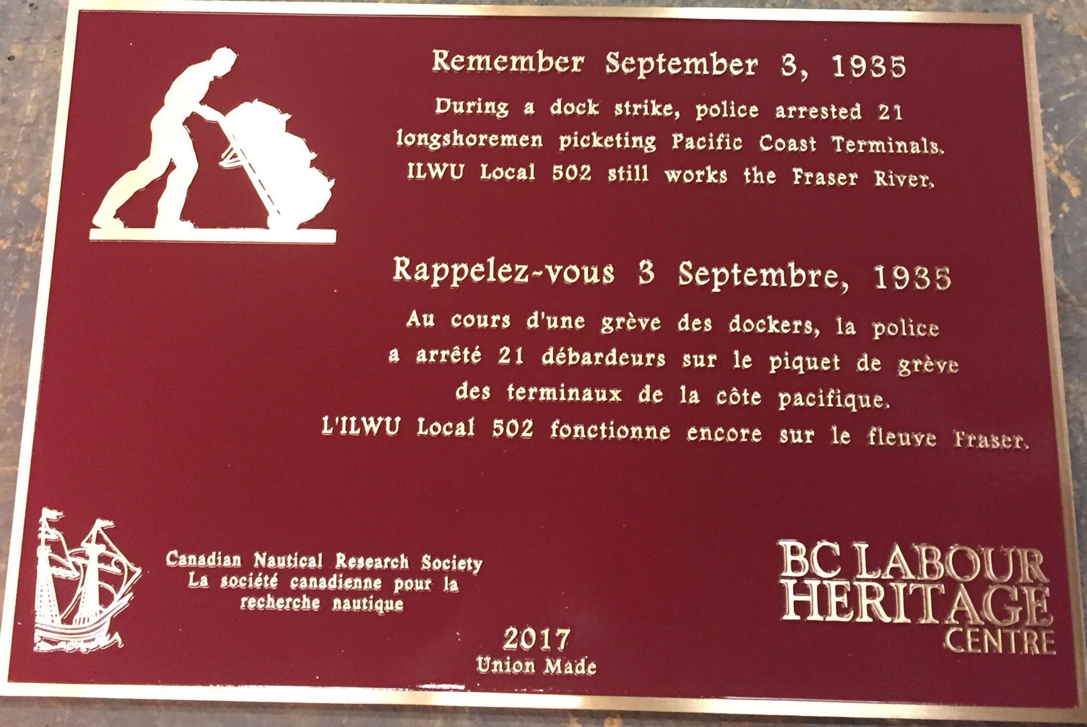 The plaque reads: Remember September 3, 1935 During a dock strike, police arrested 21 longshoremen picketing Pacific Coast Terminals. ILWU Local 502 still works the Fraser River. Rappelez-vous 3 Septembre, 1935 Au cours d'une grève des dockers, la police a arrêté 21 débardeurs sur le piquet de grève des terminaux de la côte pacifique. L'ILWU Local 502 fonctionne encore sur le fleuve Fraser. Canadian Nautical Research Society La société canadienne pour la recherche nautique BC Labour Heritage Centre 2017 Union Made