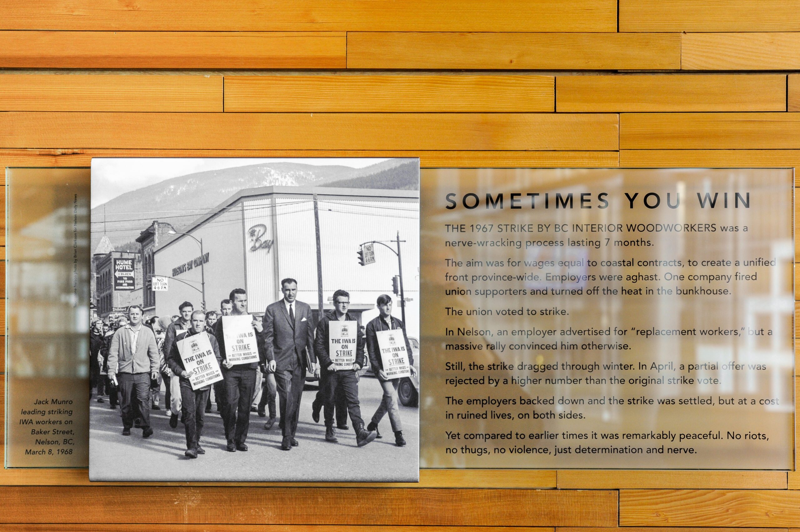The plaque reads: The 1967 strike by BC Interior Woodworkers was a nerve-wracking process lasting seven months. The aim was for wages equal to coastal contracts, to create a unified front province-wide. Employers were aghast. One company fired union supporters and turned off the heat in the bunkhouse. The union voted to strike. In Nelson, an employer advertised for “replacement workers,” but a massive rally convinced him otherwise. Still, the strike dragged through the winter. In April, a partial offer was rejected by a higher number than the original strike vote. The employers backed down and the strike was settled, but at a cost in ruined lives on both sides. Yet compared to earlier times, it was remarkably peaceful. No riots, no thugs, no violence, just determination and nerve.