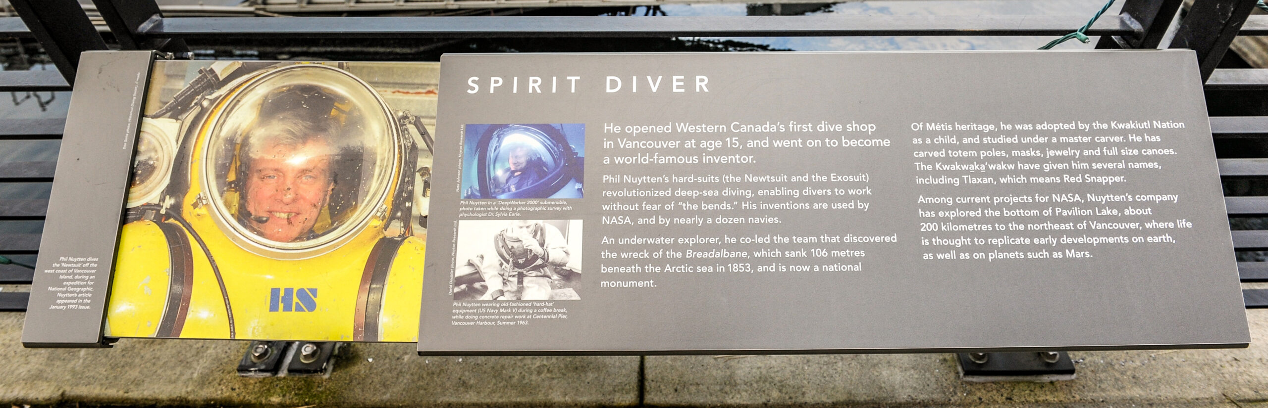 The plaque reads: He opened Western Canada’s first dive shop in Vancouver at age 15, and went on to become a world-famous inventor. Phil Nuytten’s hard-suits (the Newtsuit and the Exosuit) revolutionized deep-sea diving, enabling divers to work without fear of “the bends.” His inventions are used by NASA, and by nearly a dozen navies. An underwater explorer, he co-led the team that discovered the wreck of the Breadalbane, which sank 106 metres beneath the Arctic sea in 1853, and is now a national monument. Of Métis heritage, he was adopted by the Kwakiutl Nation as a child, and studied under a master carver. He has carved totem poles, masks, jewelry and full size canoes. The Kwakwaka’wakw have given him several names, including Tlaxan, which means Red Snapper. Among current projects for NASA, Nuytten’s company has explored the bottom of Pavilion Lake, about 200 kilometres to the northeast of Vancouver, where life is thought to replicate early developments on Earth, as well as on planets such as Mars.