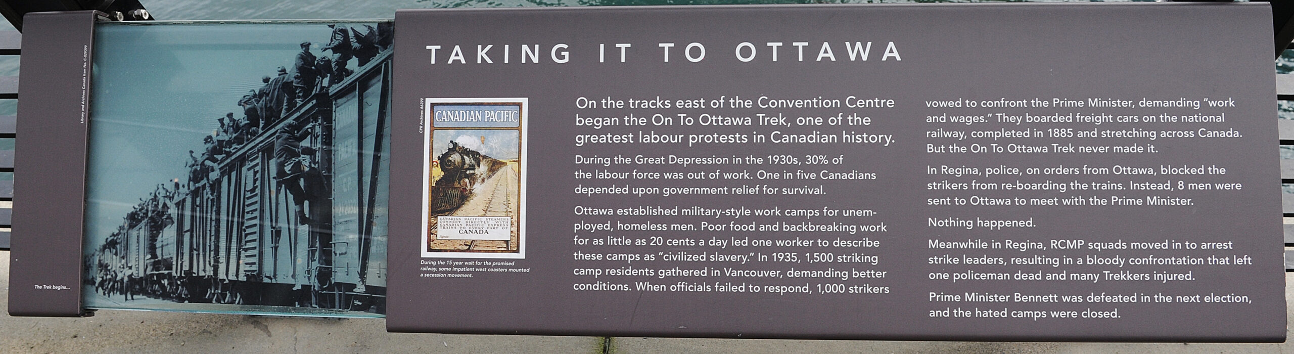 The plaque reads: On the tracks east of the Convention Centre began the On To Ottawa Trek, one of the greatest labour protests in Canadian history. During the Great Depression in the 1930s, 30% of the labour force was out of work. One in five Canadians depended upon government relief for survival. Ottawa established military-style work camps for unemployed, homeless men. Poor food and backbreaking work for as little as 20 cents a day led one worker to describe these camps as “civilized slavery.” In 1935, 1,500 striking camp residents gathered in Vancouver, demanding better conditions. When officials failed to respond, 1,000 strikers vowed to confront the Prime Minister, demanding “work and wages.” They boarded freight cars on the national railway, completed in 1885 and stretching across Canada. But the On To Ottawa Trek never made it. In Regina, police, on orders from Ottawa, blocked the strikers from re-boarding the trains. Instead, 8 men were sent to Ottawa to meet with the Prime Minister. Nothing happened. Meanwhile in Regina, RCMP squads moved in to arrest strike leaders, resulting in a bloody confrontation that left one policeman dead and many Trekkers injured. Prime Minister Bennett was defeated in the next election, and the hated camps were closed.