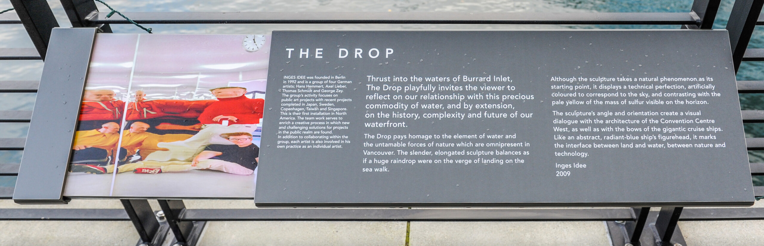 The plaque reads: Thrust into the waters of Burrard Inlet, The Drop playfully invites the viewer to reflect on our relationship with this precious commodity of water, and by extension, on the history, complexity and future of our waterfront. The Drop pays homage to the element of water and the untameable forces of nature which are omnipresent in Vancouver. The slender, elongated sculpture balances as if a huge raindrop were on the verge of landing on the sea walk. Although the sculpture takes a natural phenomenon as its starting point, it displays a technical perfection, artificially coloured to correspond to the sky, and contrasting with the pale yellow of the mass of sulfur visible on the horizon. The sculpture’s angle and orientation create a visual dialogue with the architecture of the Convention Centre West, as well as with the bows of the gigantic cruise ships. Like an abstract, radiant-blue ship’s figurehead, it marks the interface between land and water, between nature and technology. Inges Idee 2009