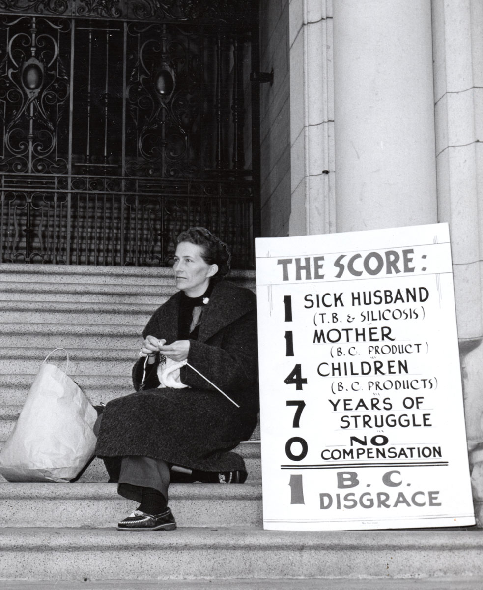 Bea Zucco sitting on the steps of the BC legislature knitting with a sign next to her that lists the score