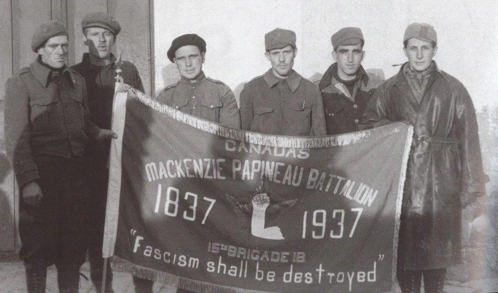 Black and white photo of 6 men holding up a banner that say: "Canadas Mackenzie Papineau Battalion"